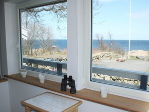 Ein Zimmer mit großem Fenster, das auf eine felsige Küstenlinie und das Meer blickt. Eine Holzablage mit zwei weißen Krügen und einem Fernglas.
