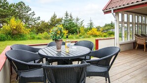 A covered outdoor patio with a round table and six wicker chairs, surrounded by a wooden deck and a glass enclosure.