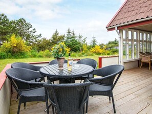 En overdækket terrasse med et rundt bord og seks fletstole, omgivet af et træterrasse og et glasindhegning.