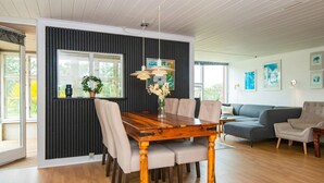 A dining area with a wooden table, chairs, and a pendant light. There is a dark wall with vertical lines, a window, and a sofa in the background.