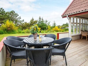 Eine überdachte Terrasse im Freien mit einem runden Tisch und sechs Korbstühlen, umgeben von einer Holzterrasse und einer Glaskonstruktion.