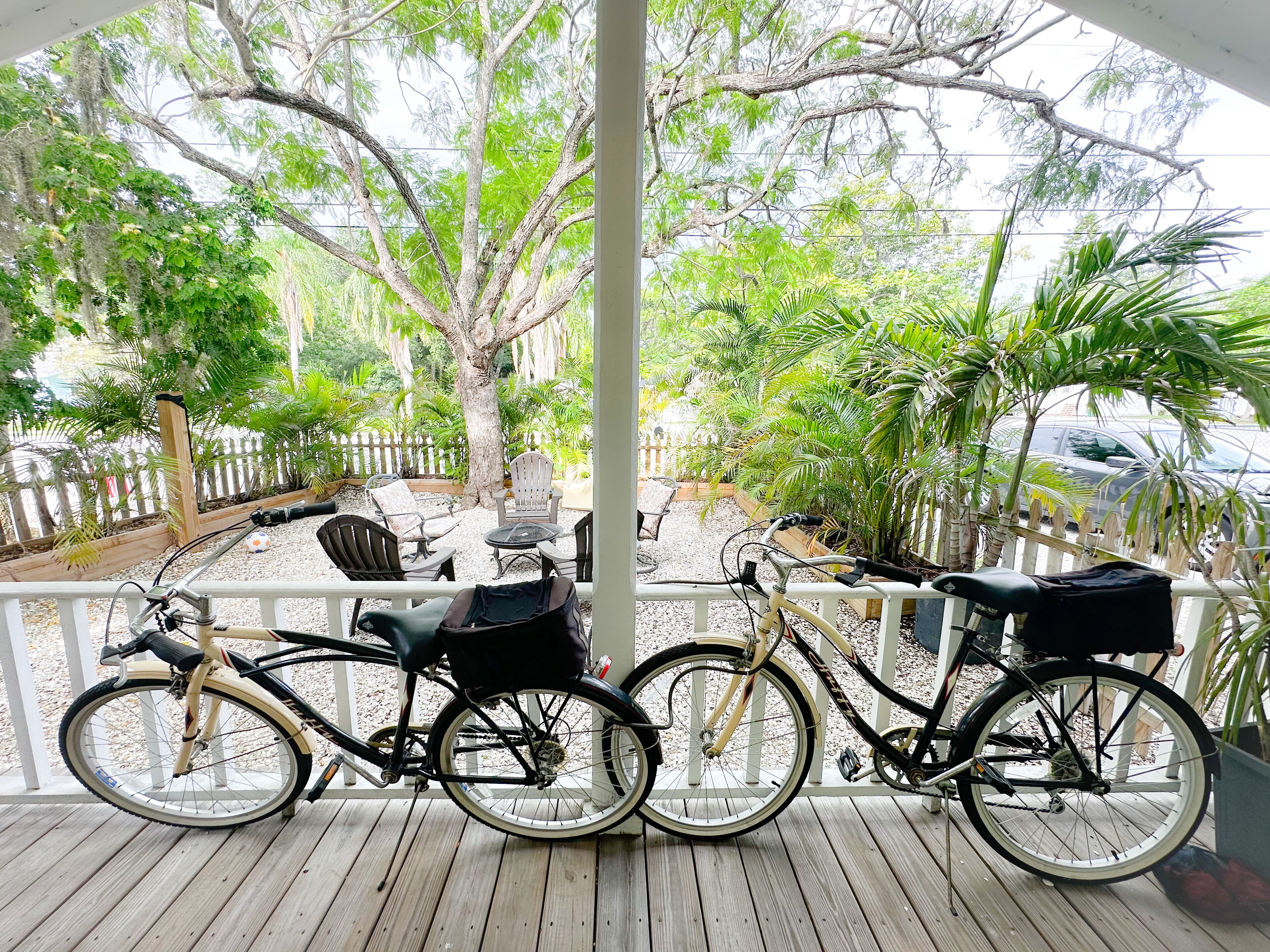 A covered porch with two bicycles parked, surrounded by lush greenery and a white fence.