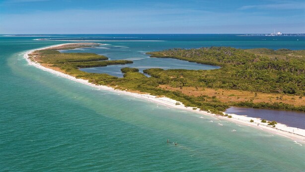 Vue aérienne d’une zone côtière avec une plage de sable, des eaux turquoise et limpides, et une végétation luxuriante.