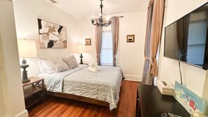 Une chambre à coucher avec un lit, deux lampes de chevet, un lustre, un téléviseur et un tableau encadré au mur.