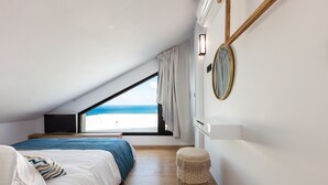 Um quarto com uma cama, um banquinho pequeno, um espelho redondo e vista para a praia.