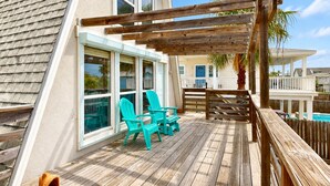 Una terraza de madera con sillas, una pérgola y vistas a una piscina y casas vecinas.