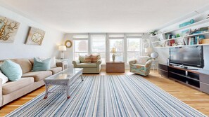 A living room with a sofa, armchair, coffee table, TV, bookshelf, and large windows.