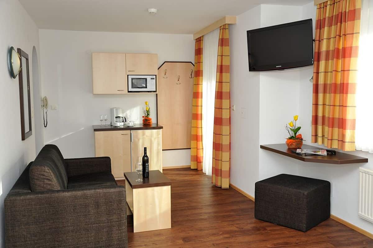 A compact living space with a sofa, a small table with a bottle and glasses, a kitchen area with a microwave, and a TV mounted on the wall.