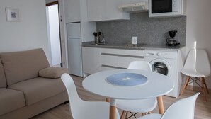 Cocina pequeña equipada con microondas, cafetera y lavadora. Hay una mesa redonda con mantel individual y sillas blancas. Un sofá color beige se encuentra en la sala.