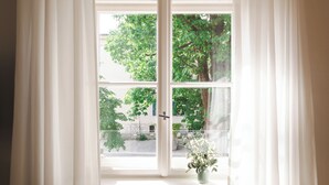 Une fenêtre avec des rideaux blancs, une plante en pot et une vue sur les arbres et les bâtiments à l’extérieur.