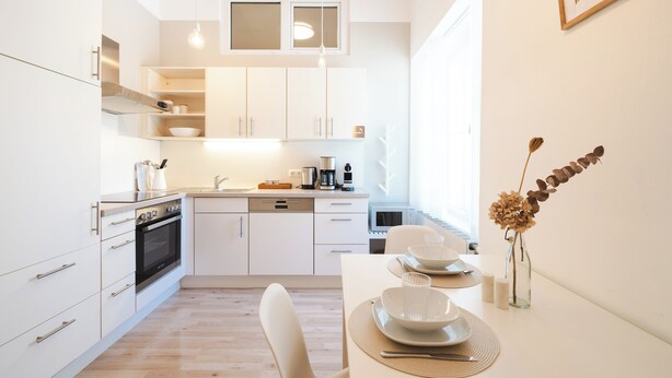 A modern kitchen with white cabinetry, a dining area with a table set for two, and a vase with dried flowers on the wall. - GrazRentals (Graz)