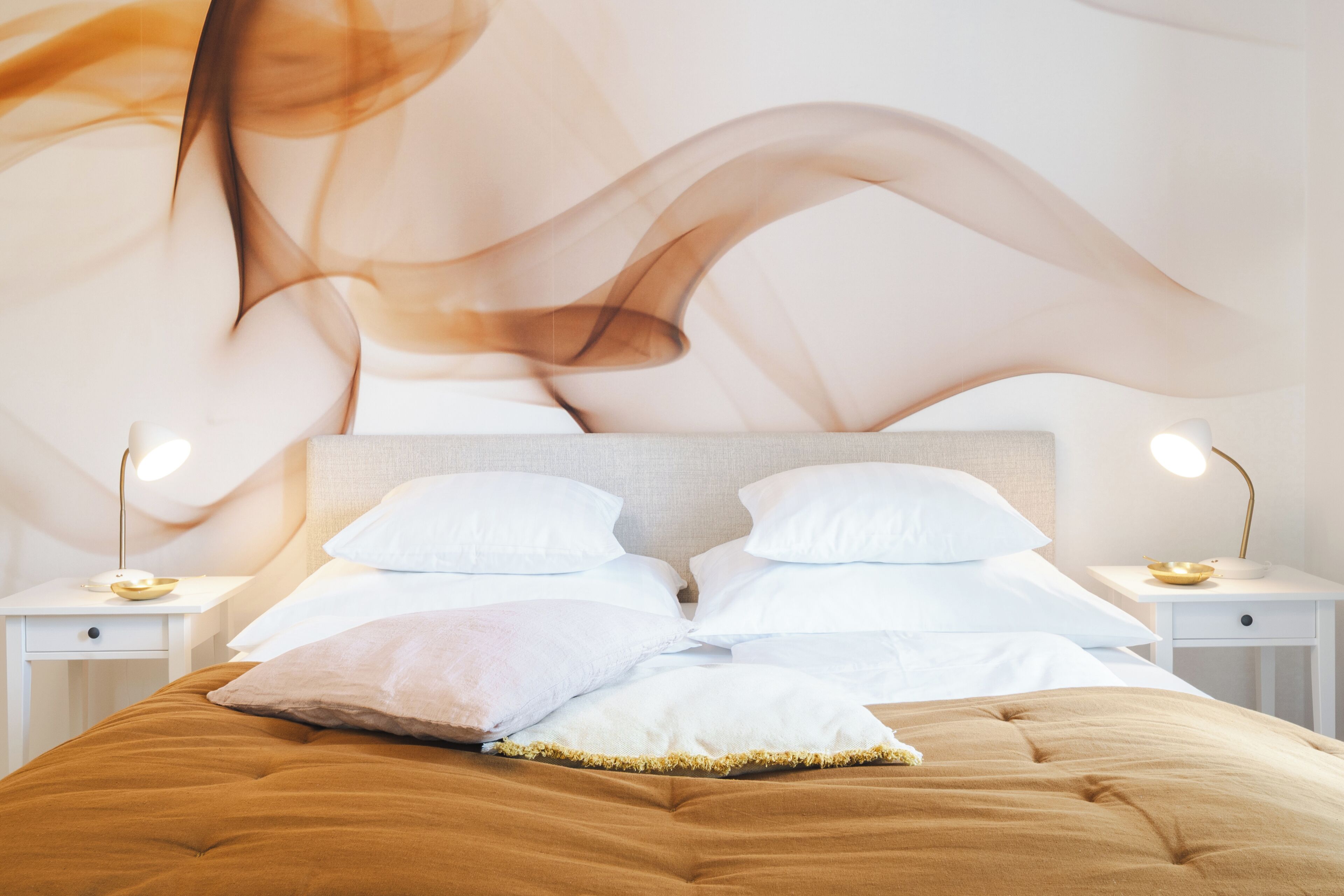Ein ordentlich bezogenes Bett mit brauner Decke, weißen Kissen und einem Kopfteil mit Wandbild. Es gibt zwei Nachttische mit Lampen.