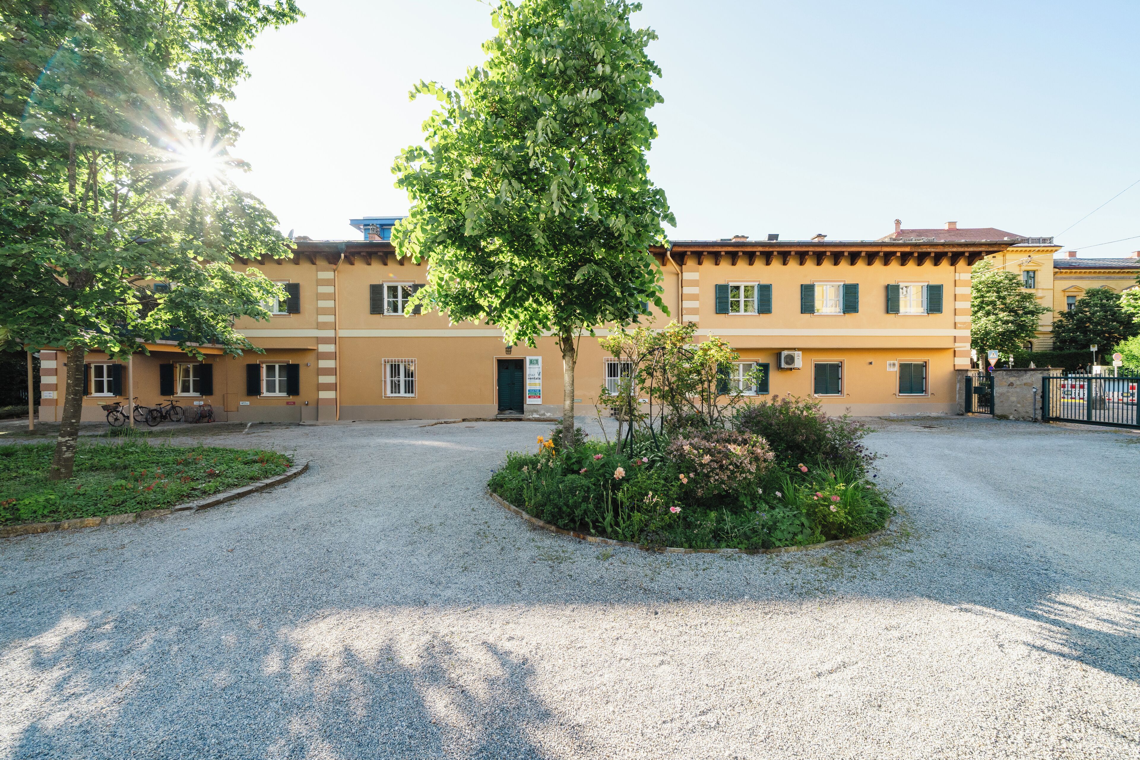A building with a gravel driveway, a garden with flowers, and a tree in front.