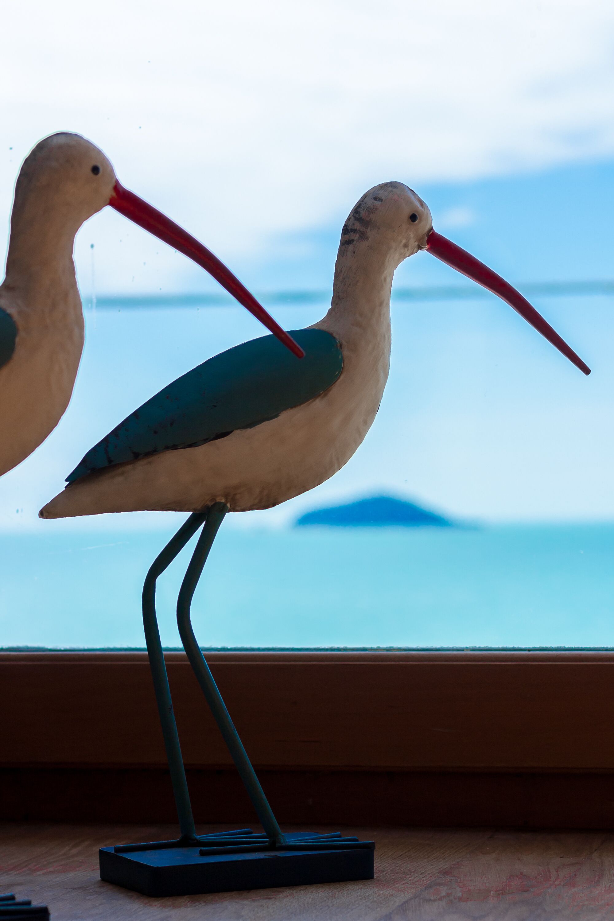 Deux sculptures en forme d’oiseaux, dotées de longs becs et de longues pattes, sont placées sur une surface en bois, près d’une fenêtre donnant sur la mer.