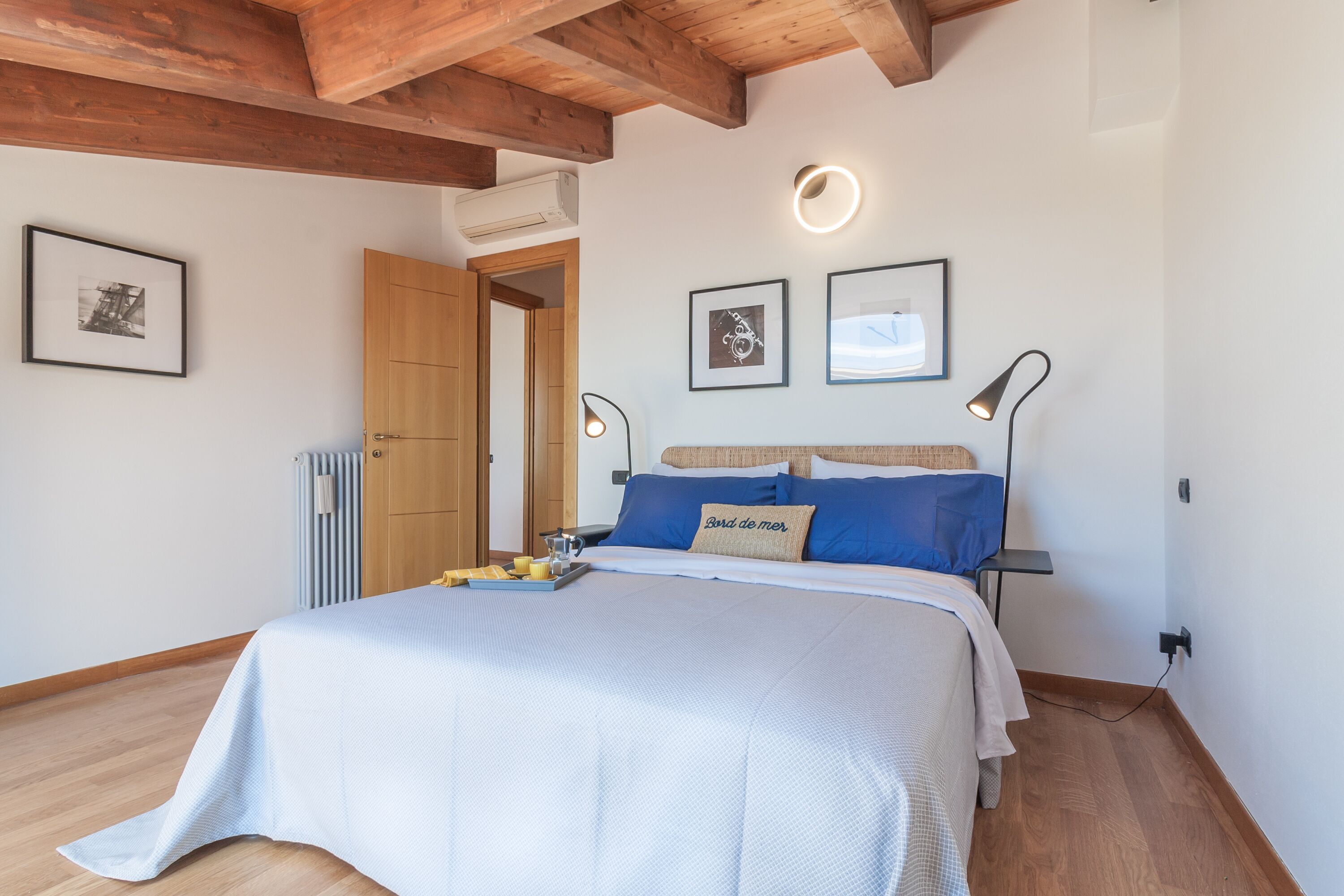 Une chambre à coucher avec un lit, des poutres en bois au plafond, un radiateur, des tableaux encadrés au mur et des lampes de chevet.