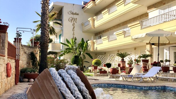 A pool area with a water feature, palm trees, and a building with balconies. - Melis Studios (Kassandra)
