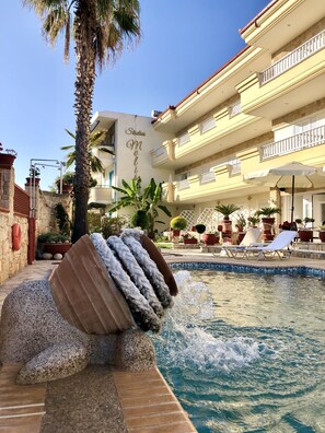 Ein Poolbereich mit Wasserspiel, Palmen und einem Gebäude mit Balkonen.