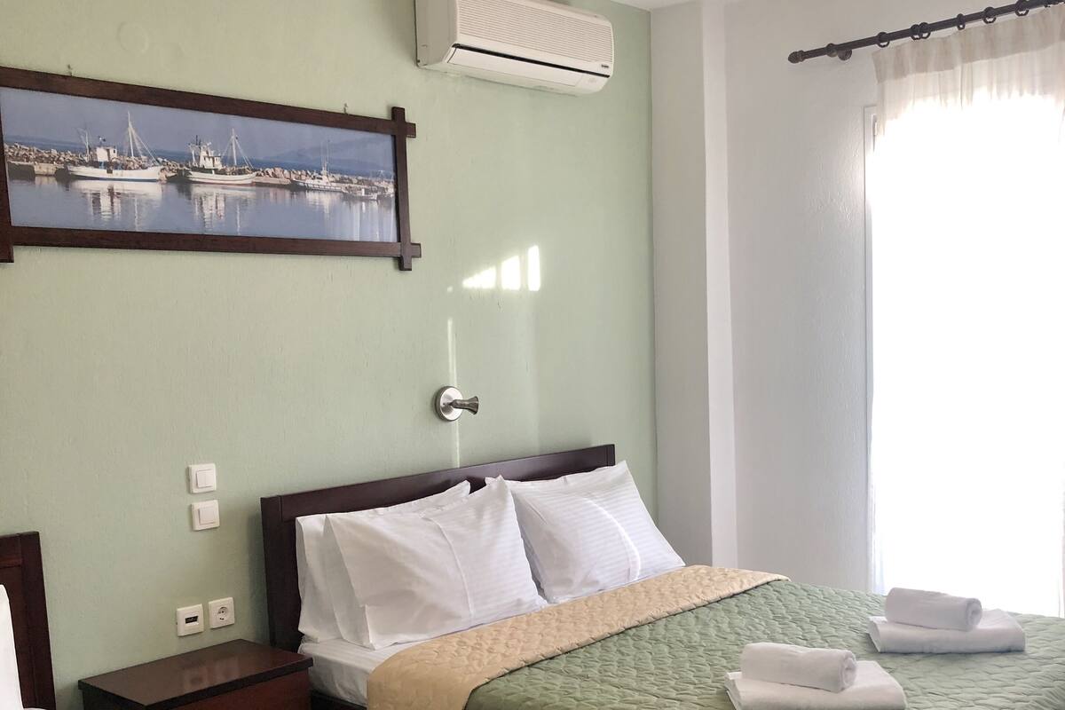 A hotel room with two beds, a painting of boats, an air conditioner, and a window with curtains.