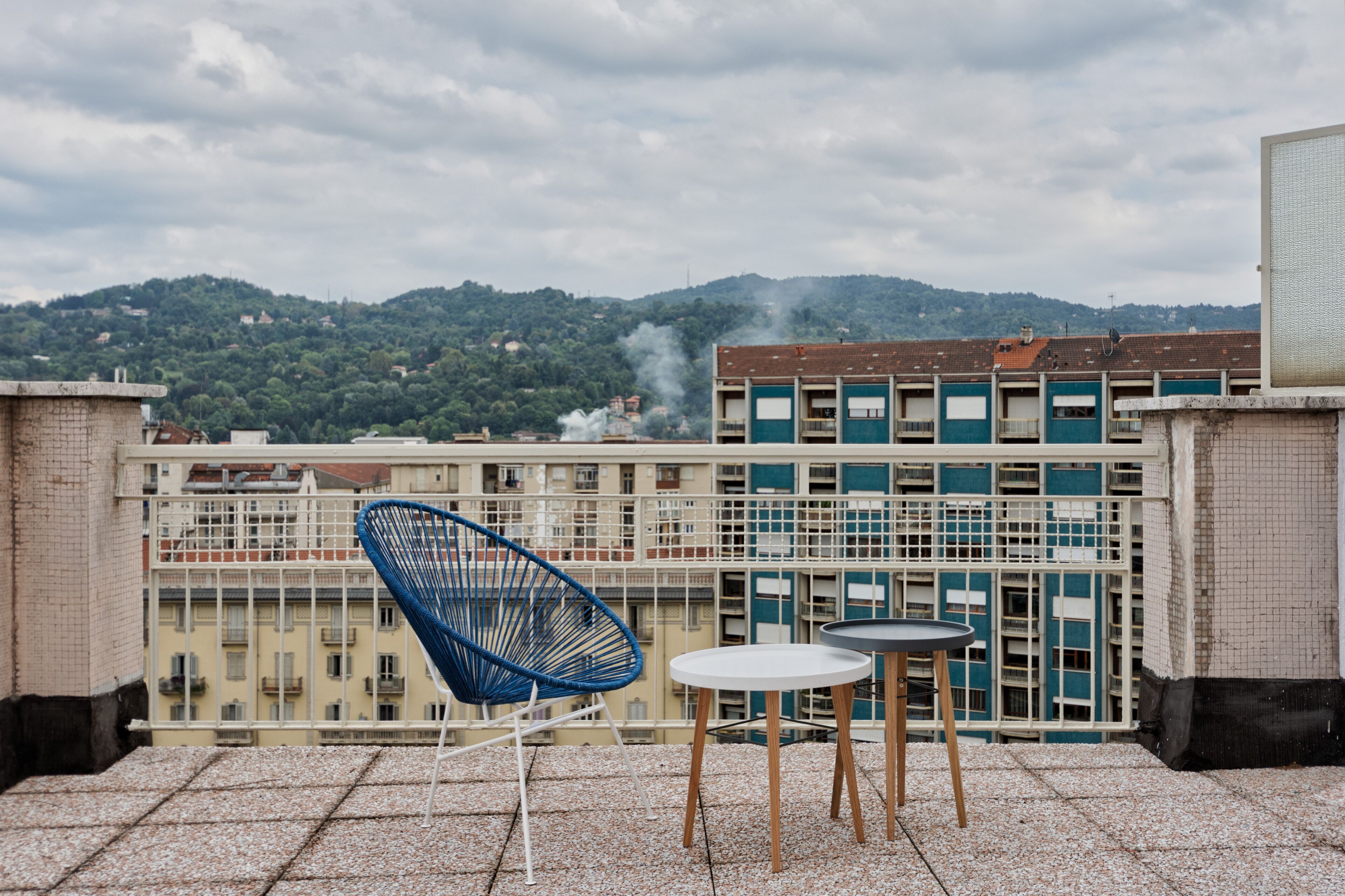 A rooftop terrace with a blue chair, a small round table, and a view of a cityscape with buildings and a hill in the background.