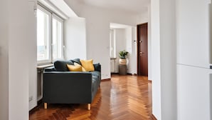 A modern living room with a dark grey sofa, wooden flooring, and a large window providing natural light. - Urban House Carducci - Attic (Turin)