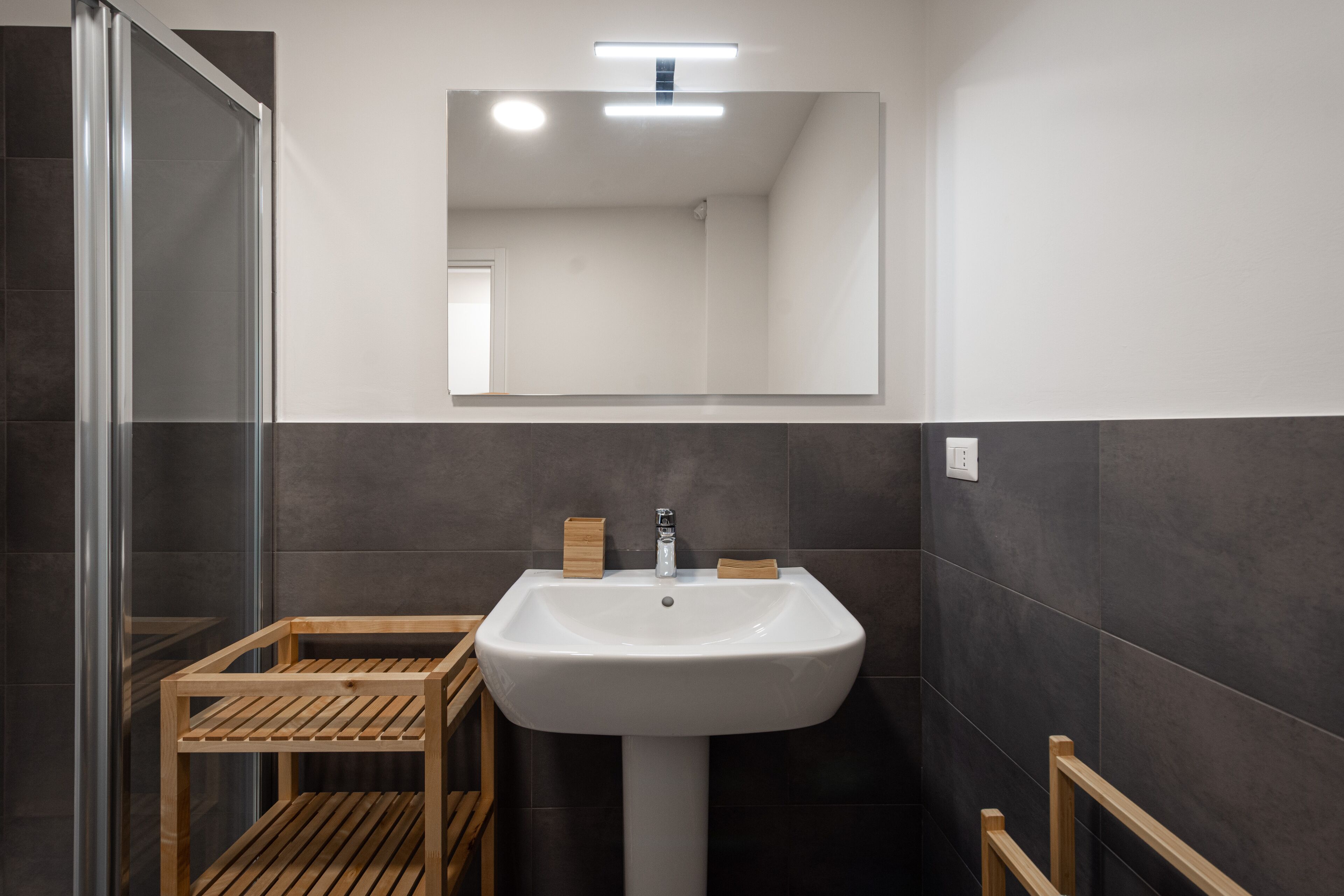 Un baño moderno con un lavamanos blanco, paredes de azulejos oscuros y un taburete de madera.