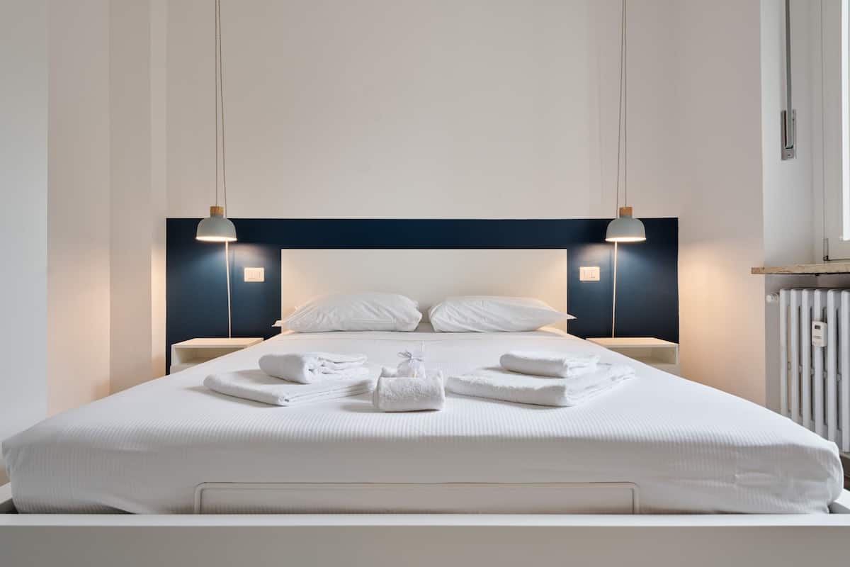 A neatly made bed with white linens and towels, two bedside tables, and pendant lights.