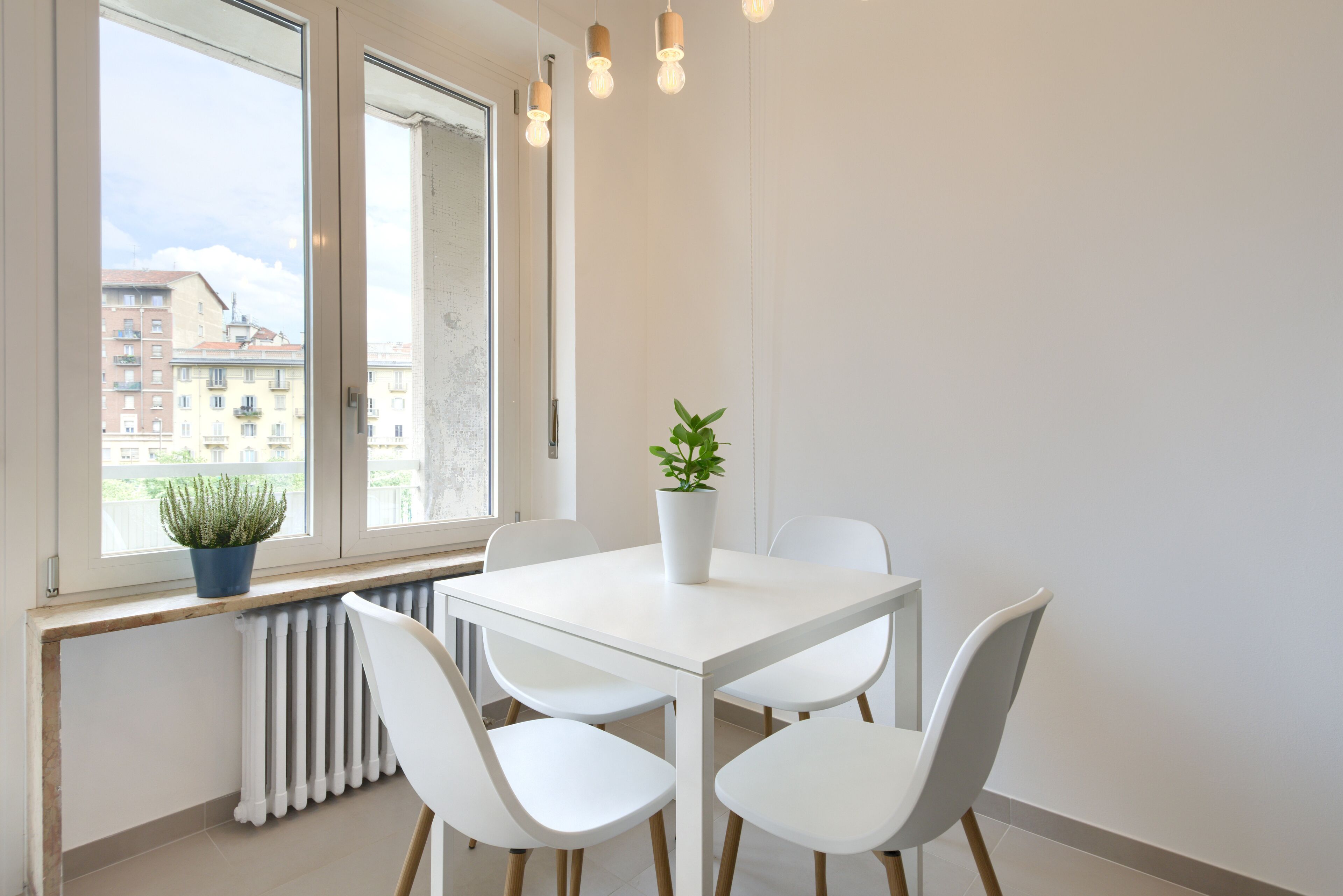 A modern dining area with a white table and chairs, a potted plant, and a view of buildings through large windows.