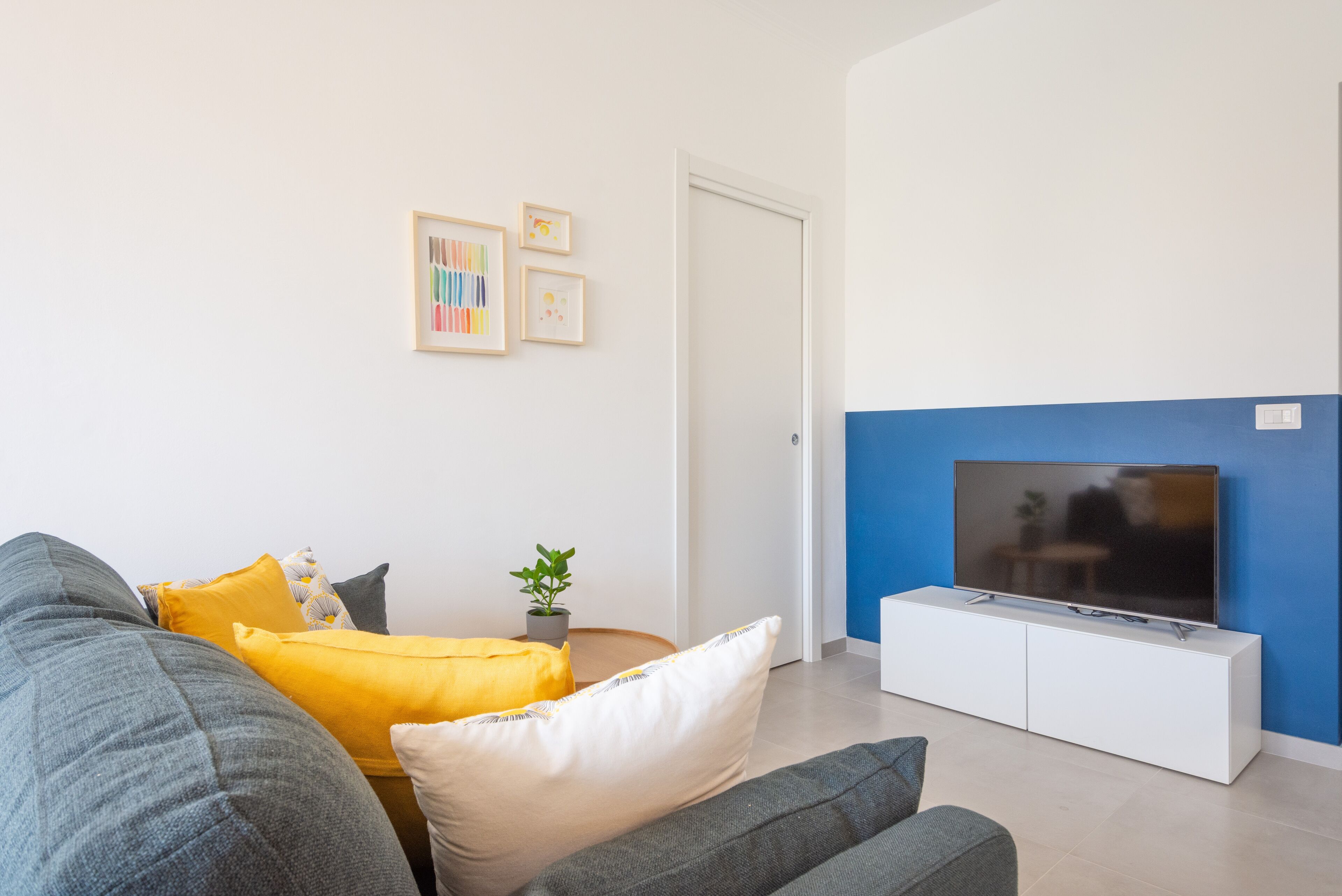 Un salon moderne avec un canapé gris, des coussins jaunes et blancs, une petite plante et un téléviseur posé sur un meuble blanc, le tout adossé à un mur bleu qui sert de couleur d’accent.