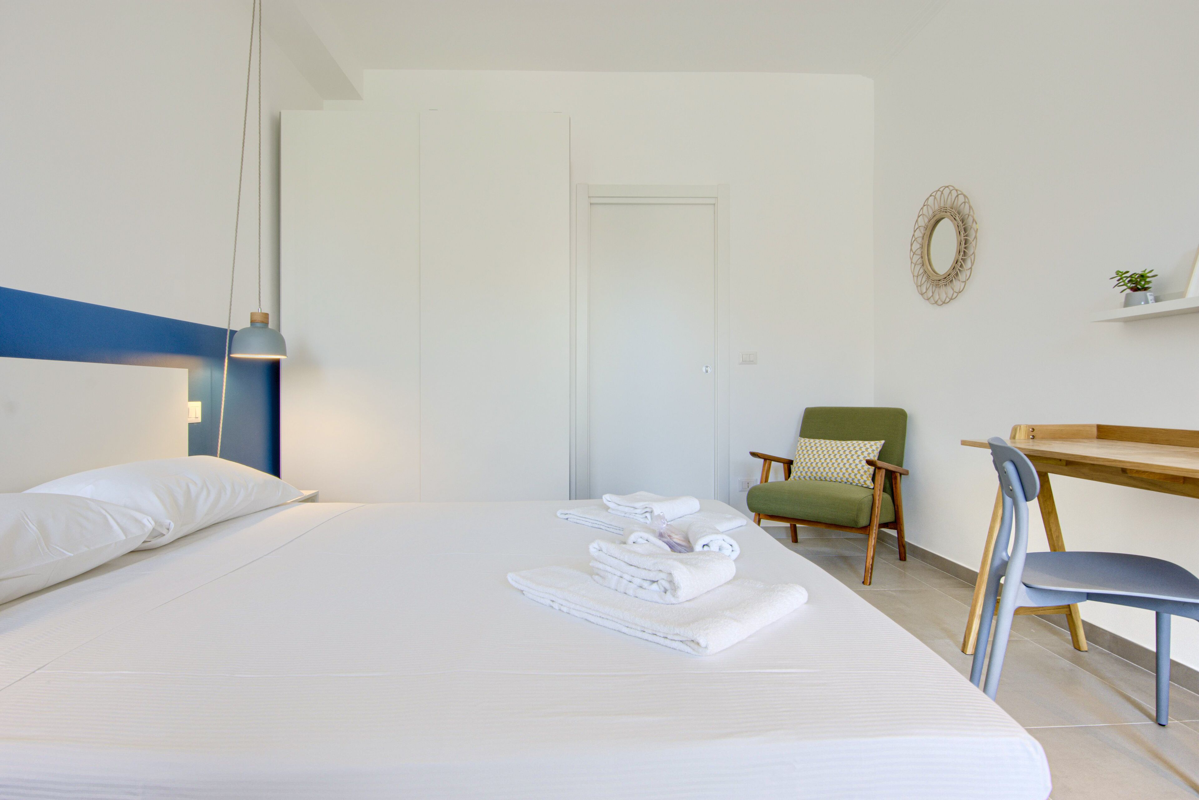 Una cama bien hecha con sábanas y toallas blancas, una silla verde, un escritorio de madera y un espejo adosado a la pared.