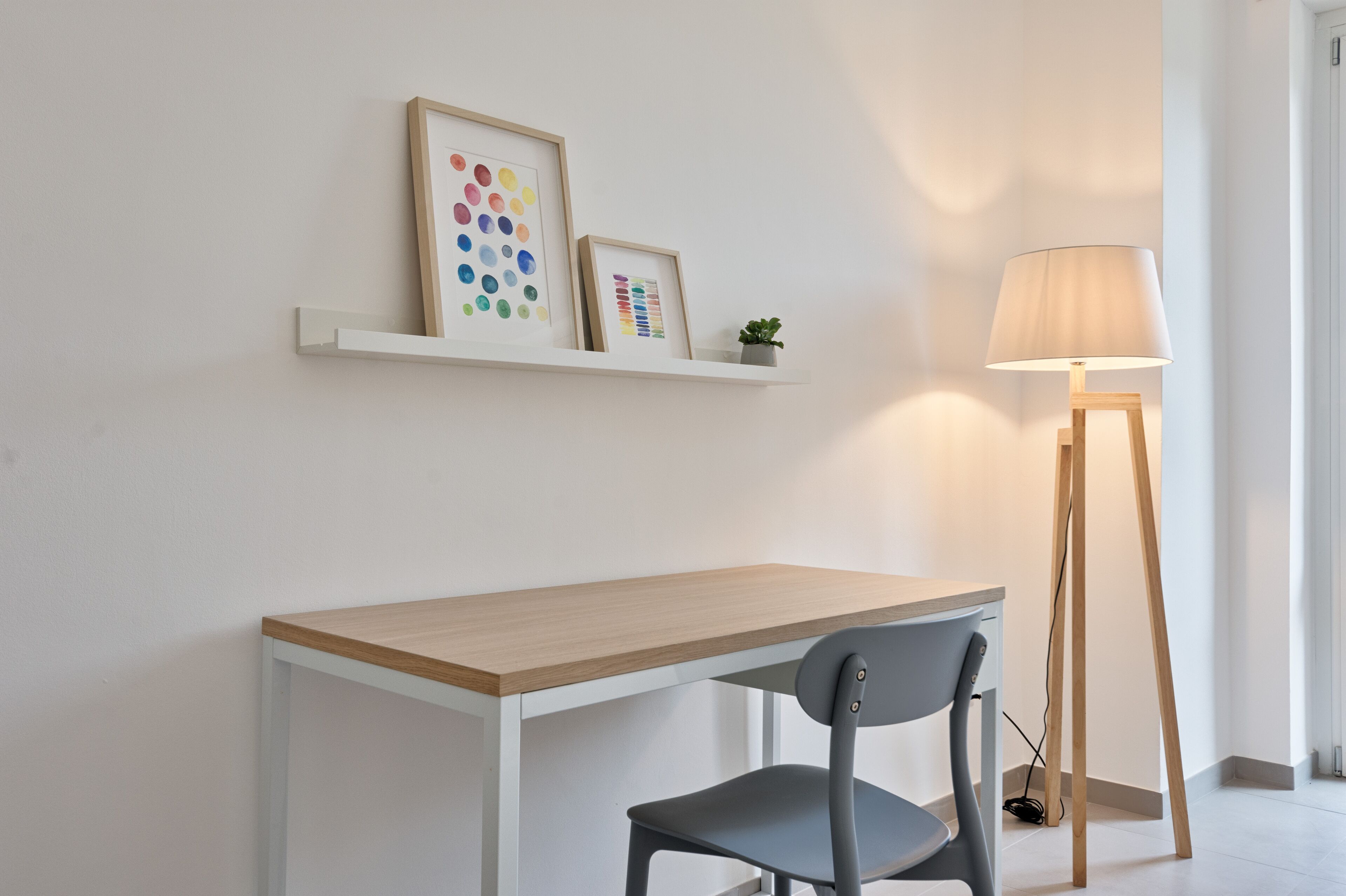 Une pièce au décor minimaliste, avec un bureau en bois, une chaise grise, une étagère blanche ornée de cadres photos et un lampadaire.