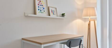 Une pièce au décor minimaliste, avec un bureau en bois, une chaise grise, une étagère blanche ornée de cadres photos et un lampadaire.