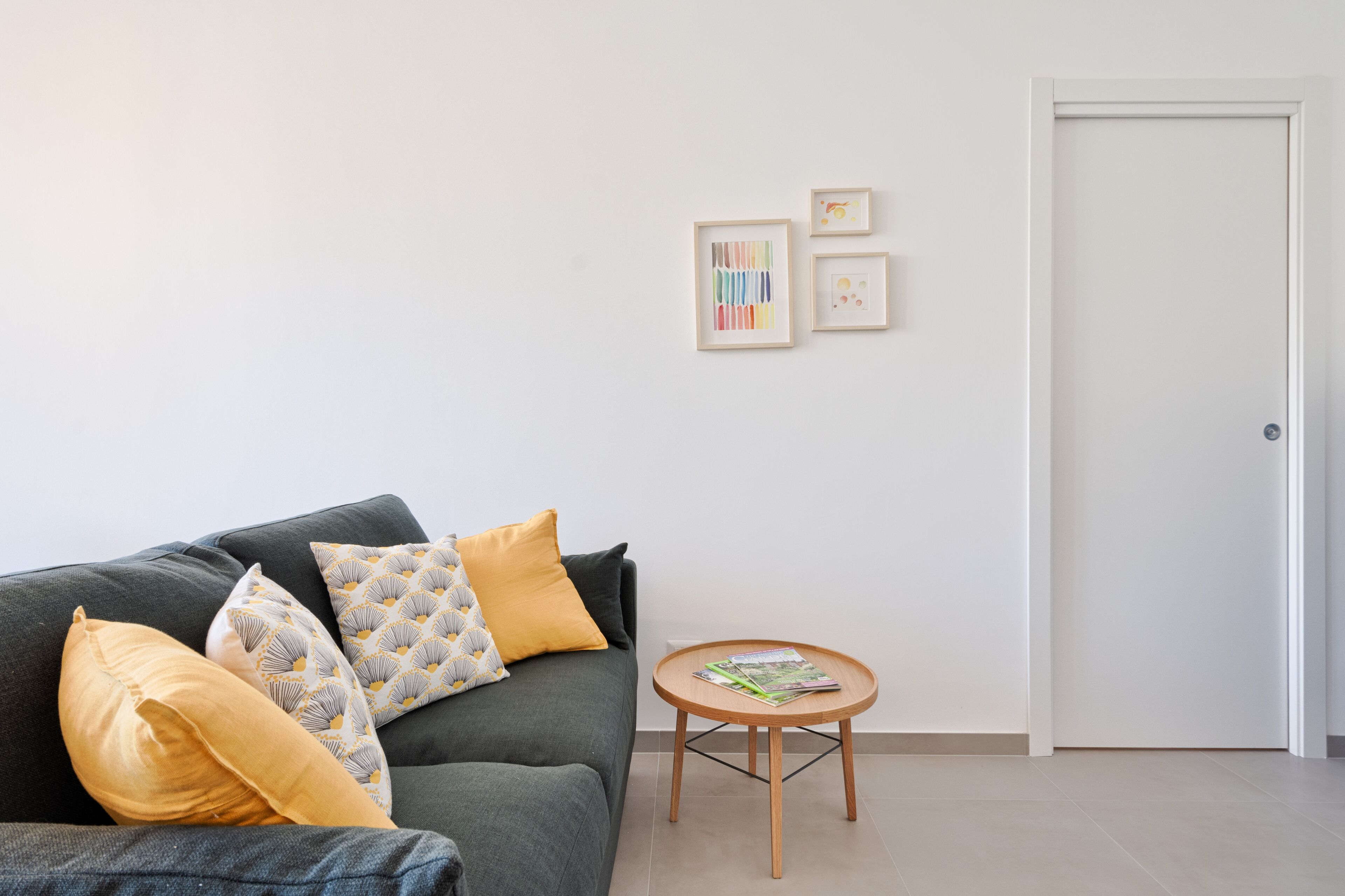 A living room with a grey sofa, yellow and patterned cushions, a small wooden side table with magazines, and a white door.