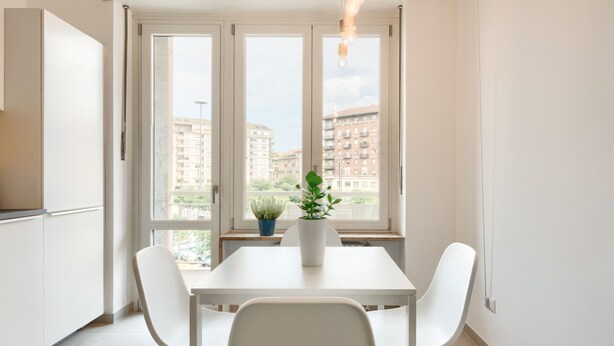 A modern kitchen with white cabinets, a white table, and chairs. There is a potted plant on the windowsill. - Urban House Carducci - Apartments (Turin)