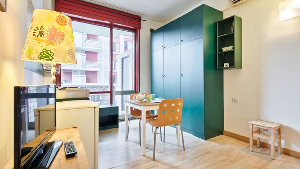 Et lille, lyst værelse med trægulv, et skrivebord med lampe, en stol, et lille bord og en reol.