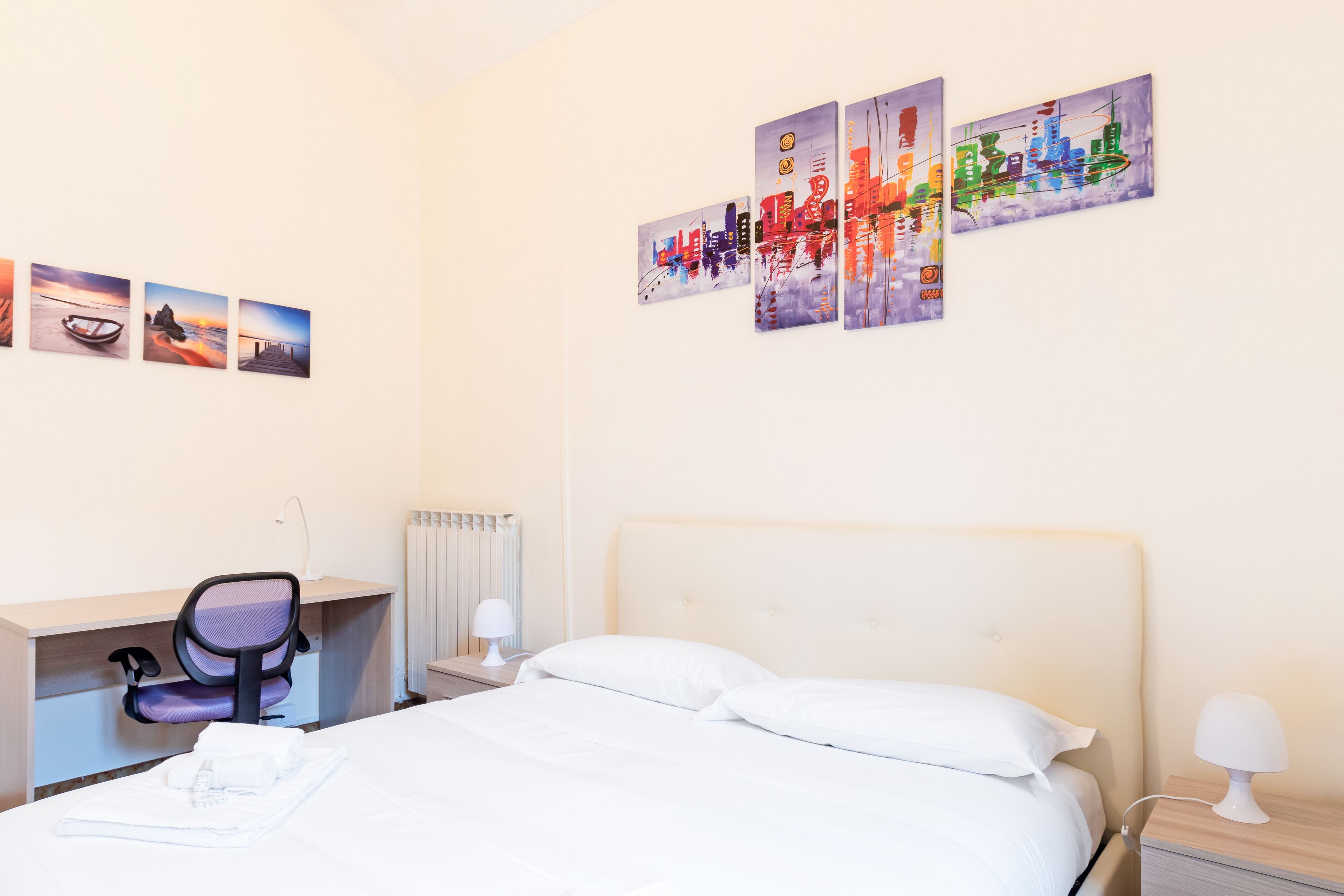 Ein Hotelzimmer mit einem Bett, einem Schreibtisch, einem Stuhl und einer Wand mit vier Kunstwerken.