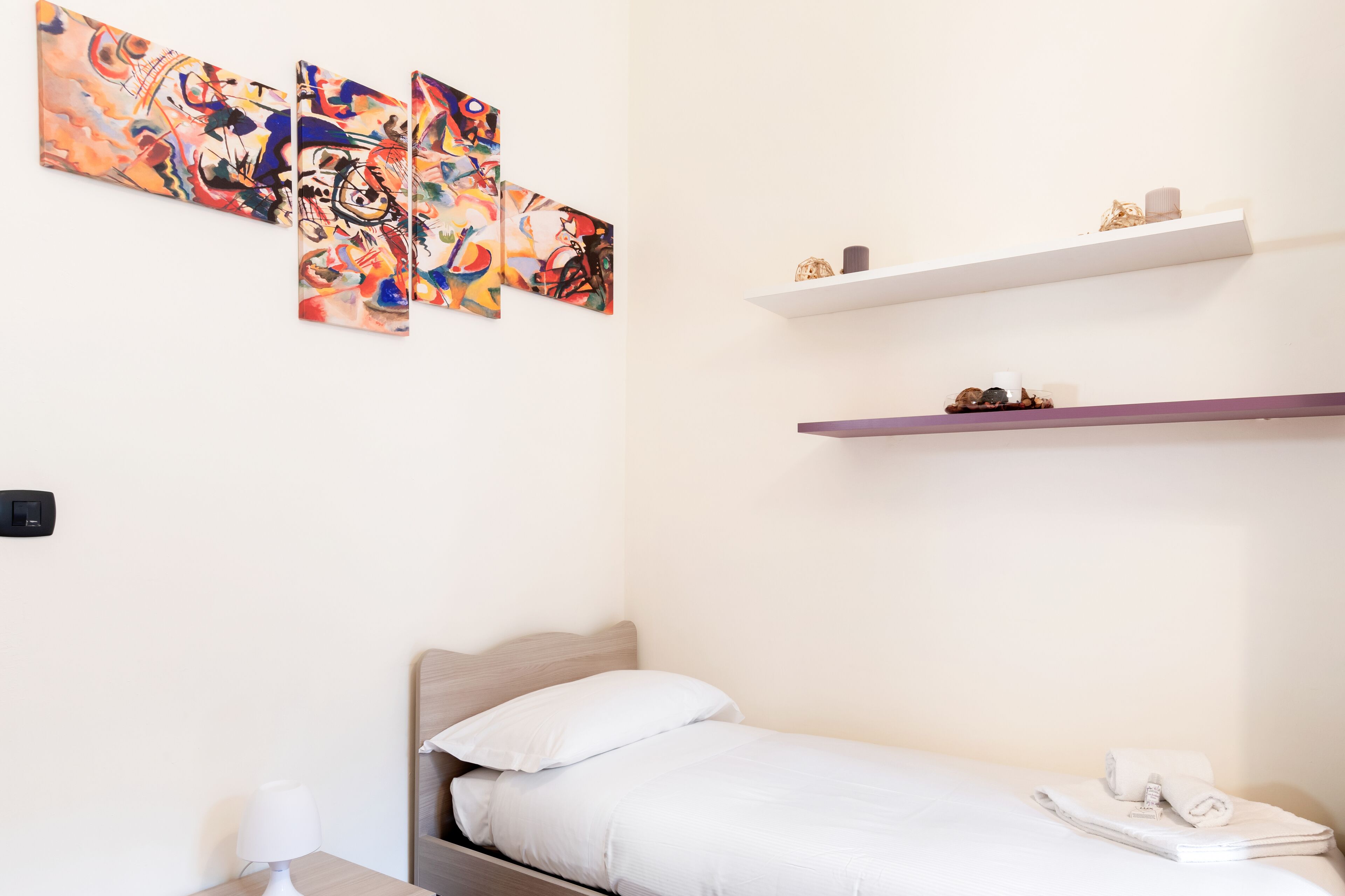 A bedroom with a bed, a nightstand, a lamp, a wall-mounted shelf with decorative items, and abstract art on the wall.