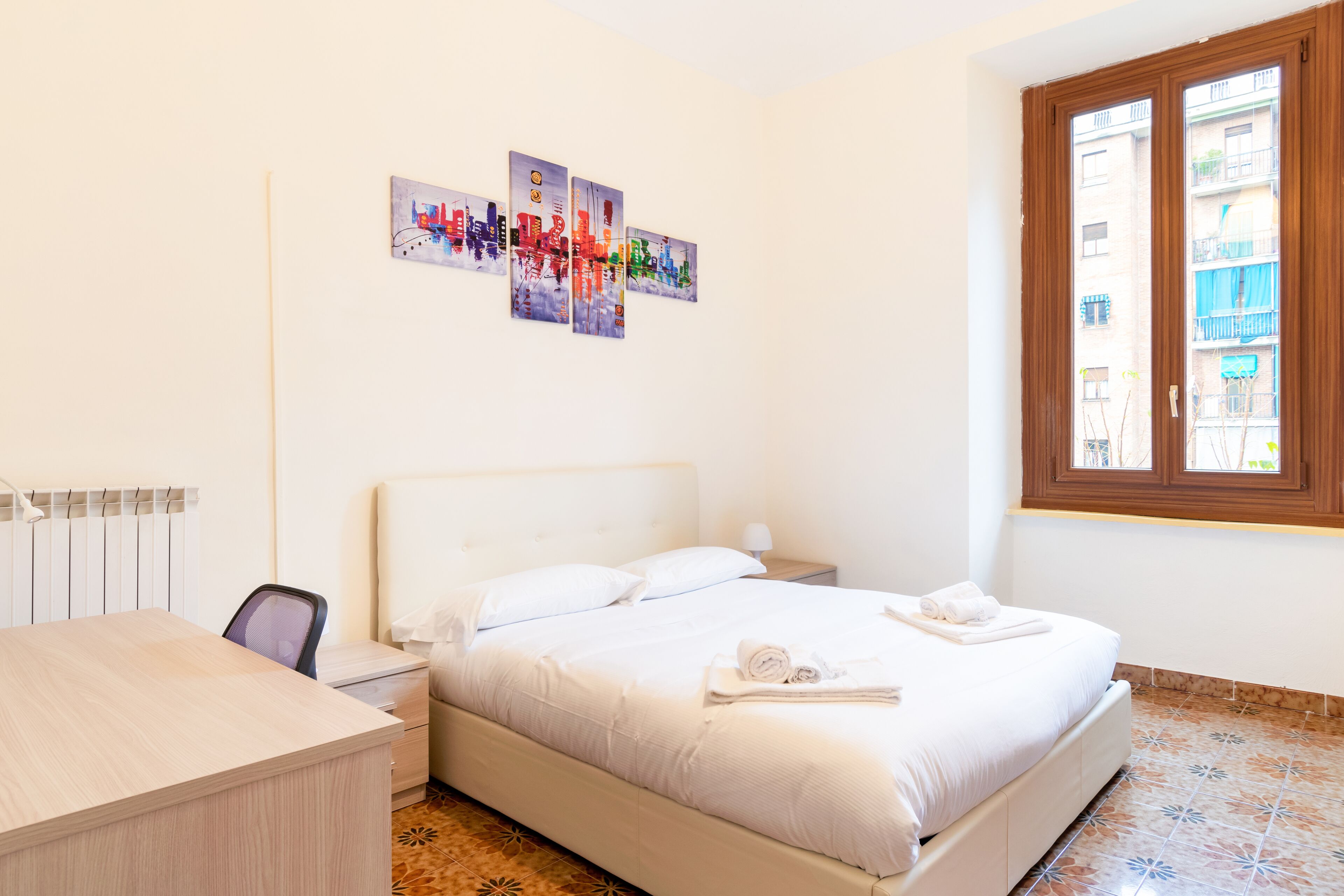 A hotel room with a bed, a desk, a chair, a radiator, and a window with a view of buildings.