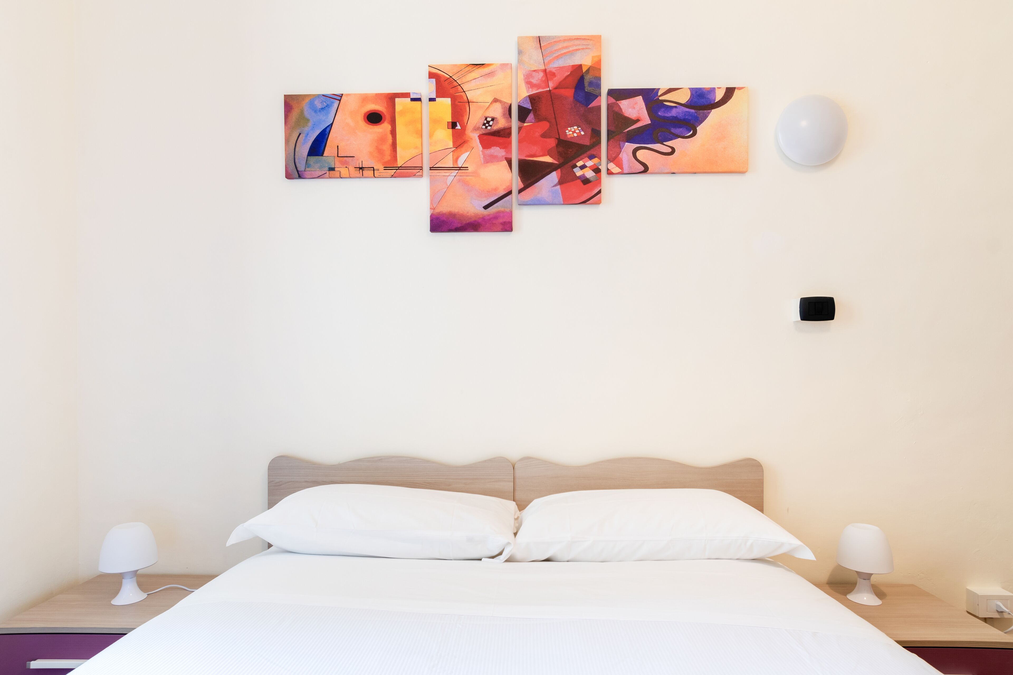 A hotel room with a bed, two bedside tables, a wall-mounted light, and a multi-panel artwork.