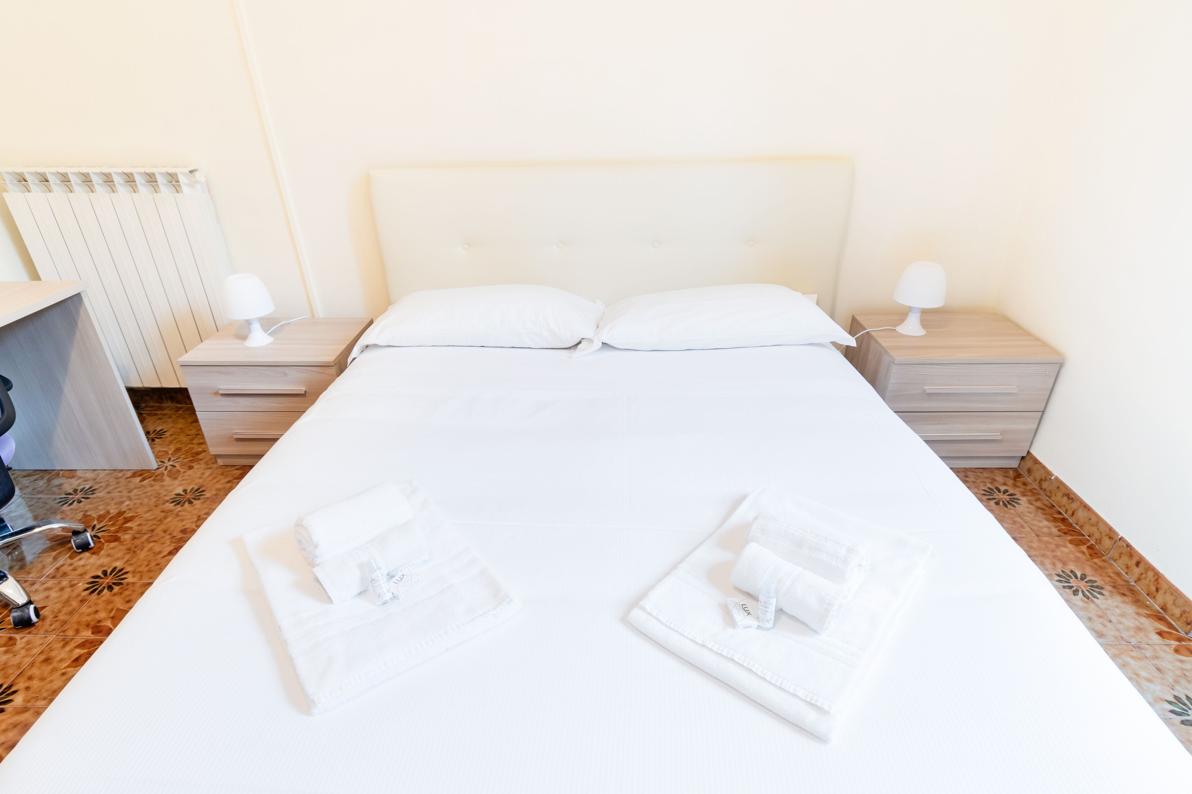A neatly made bed with white linens and two pillows, a bedside table with a lamp, and a chair with a tray.