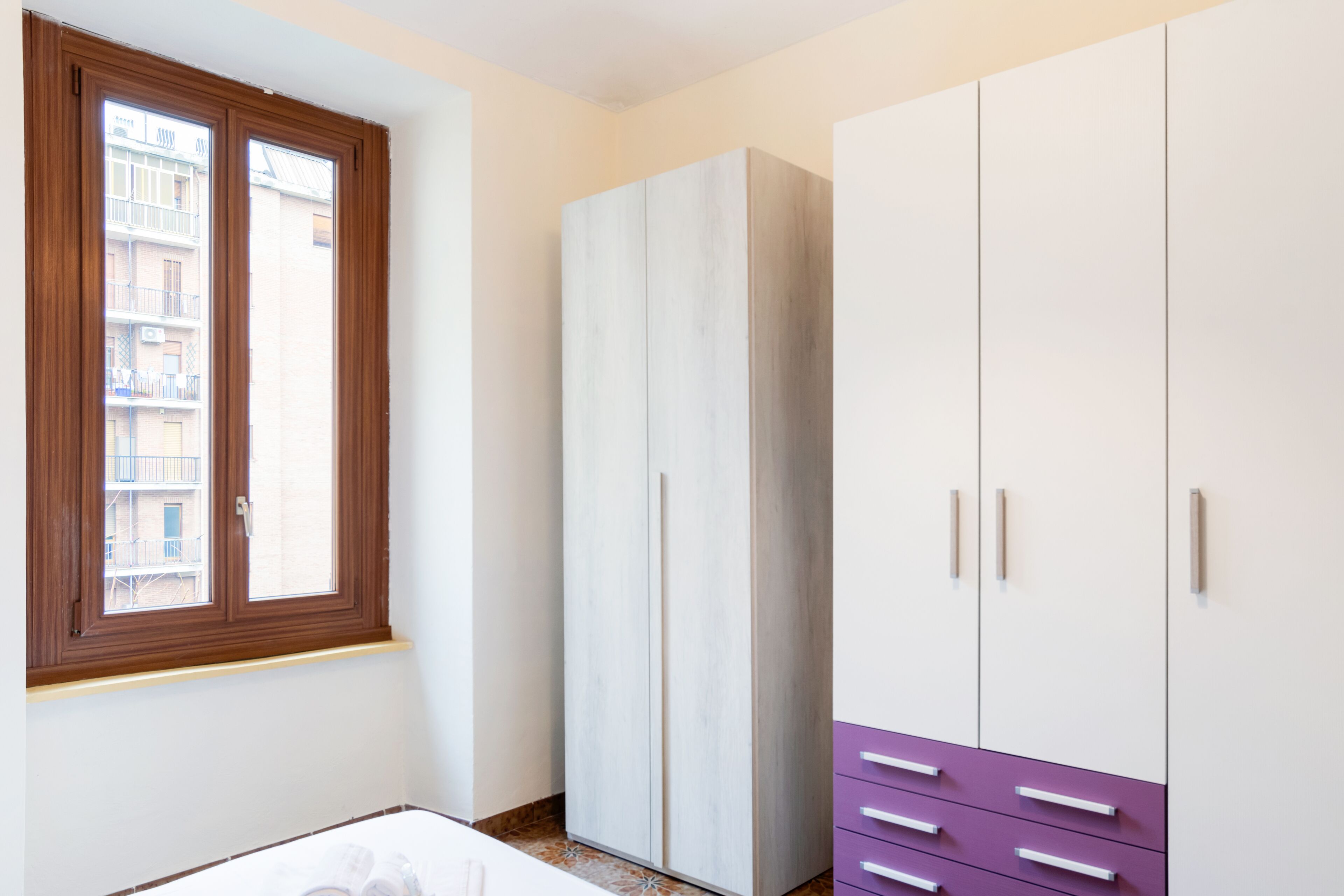 Une chambre avec une grande armoire, une fenêtre donnant sur un immeuble et un lit.