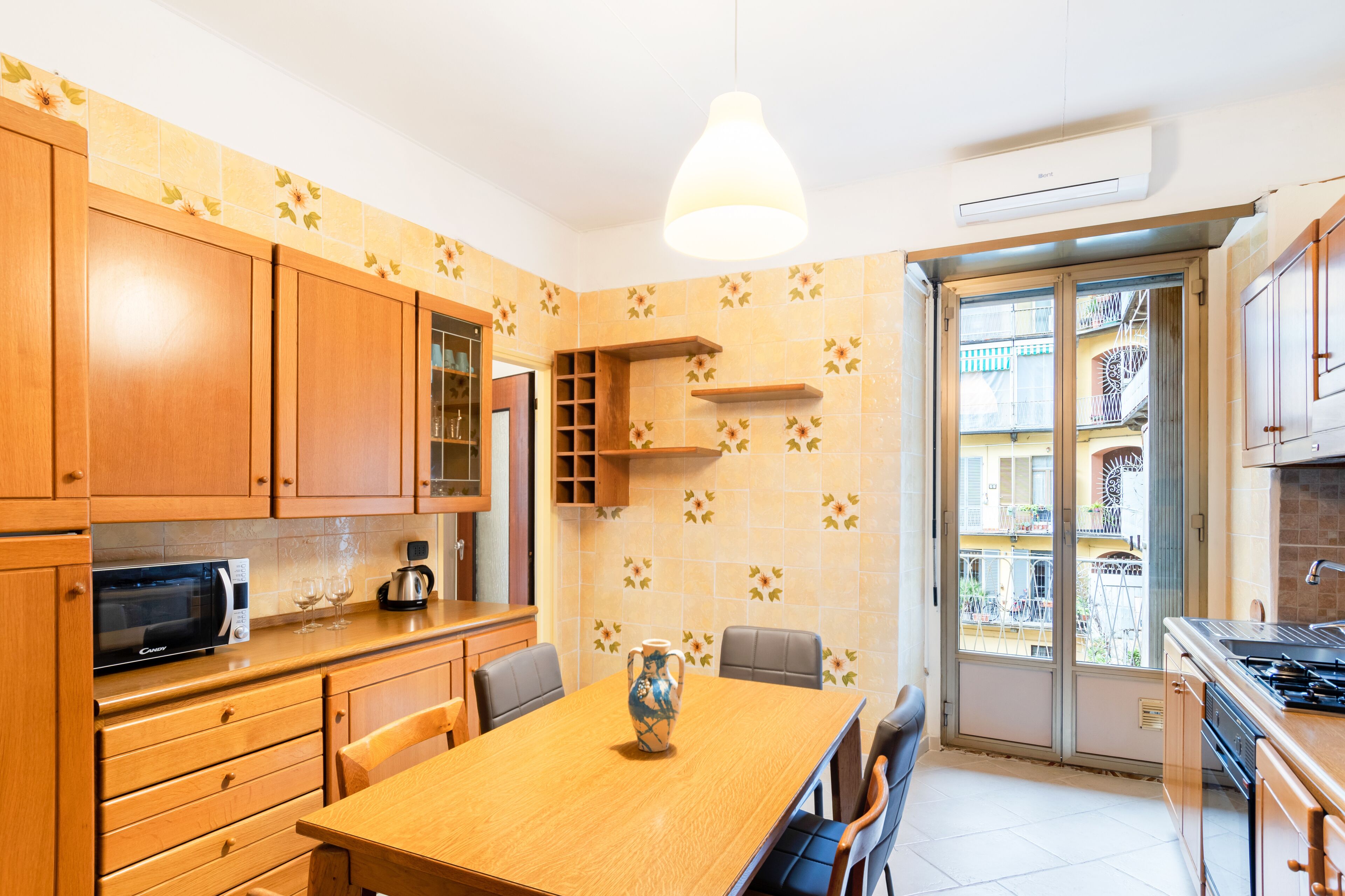 Een moderne keuken met houten kastjes, een magnetron, een eettafel met stoelen en een groot raam met uitzicht op een balkon.