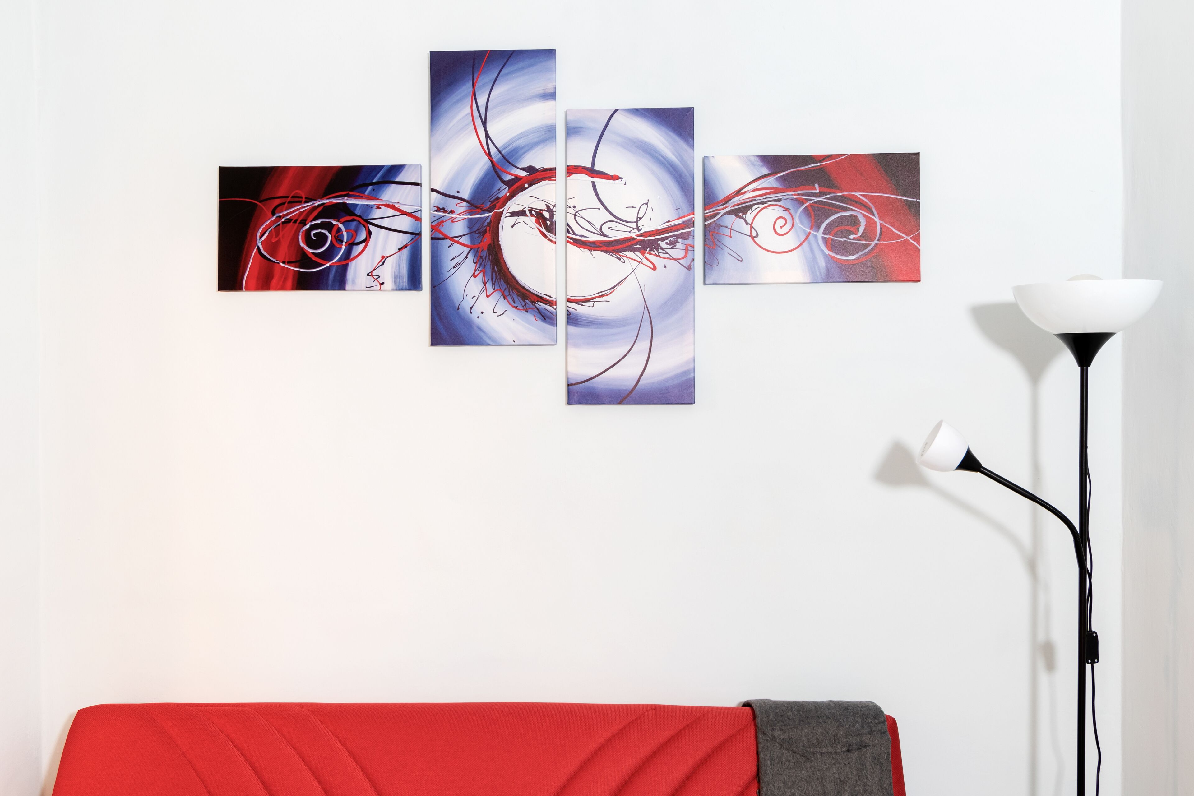 A room with a red sofa, a white floor lamp, and a wall with three abstract paintings.