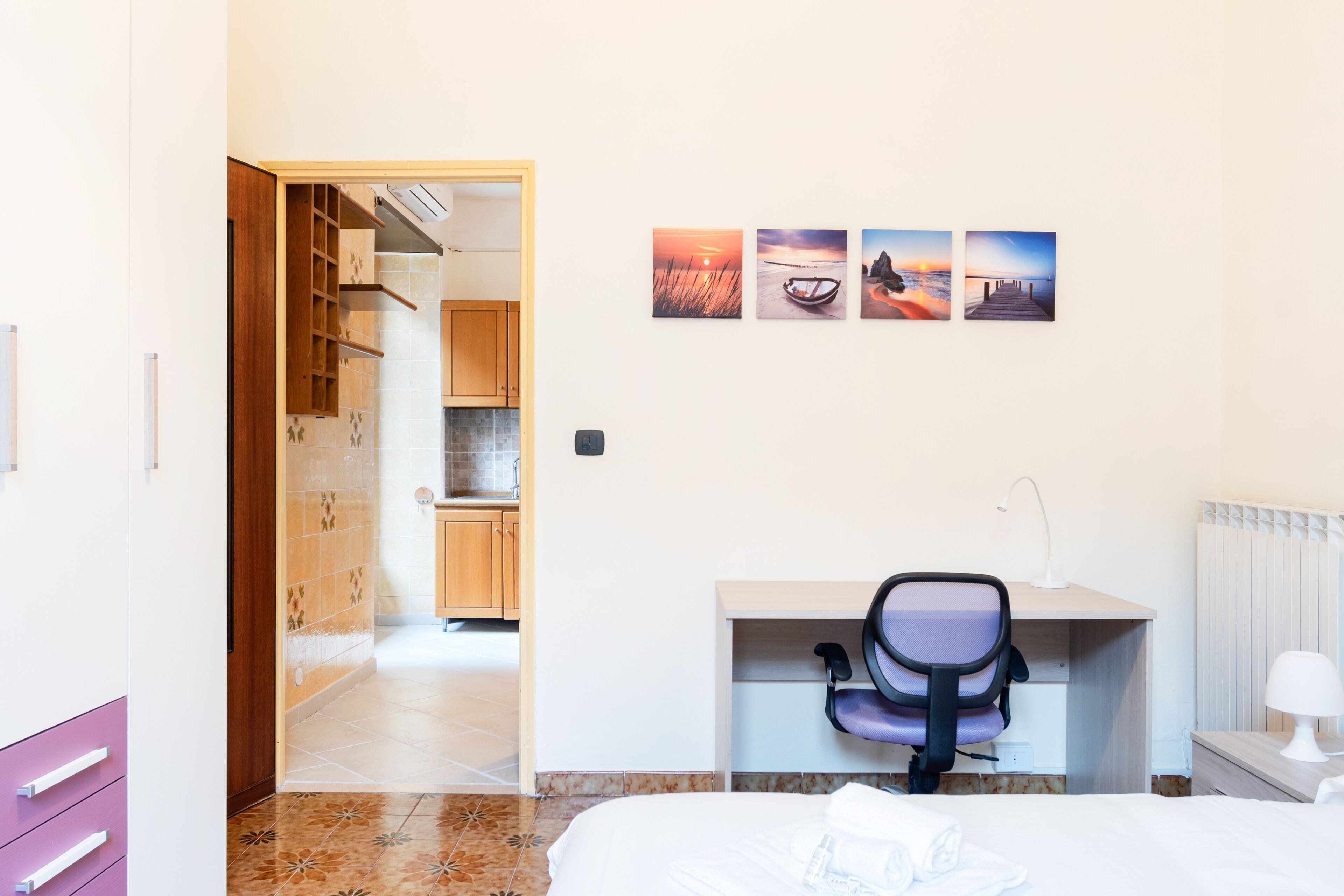 Quarto de hotel com uma cama, uma secretária com uma cadeira, um radiador e uma parede com quatro quadros.