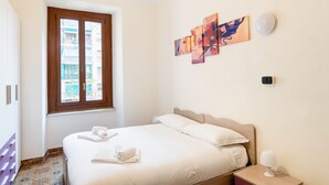 A hotel room with a bed, a window, artwork on the wall, and a bedside table with a lamp.