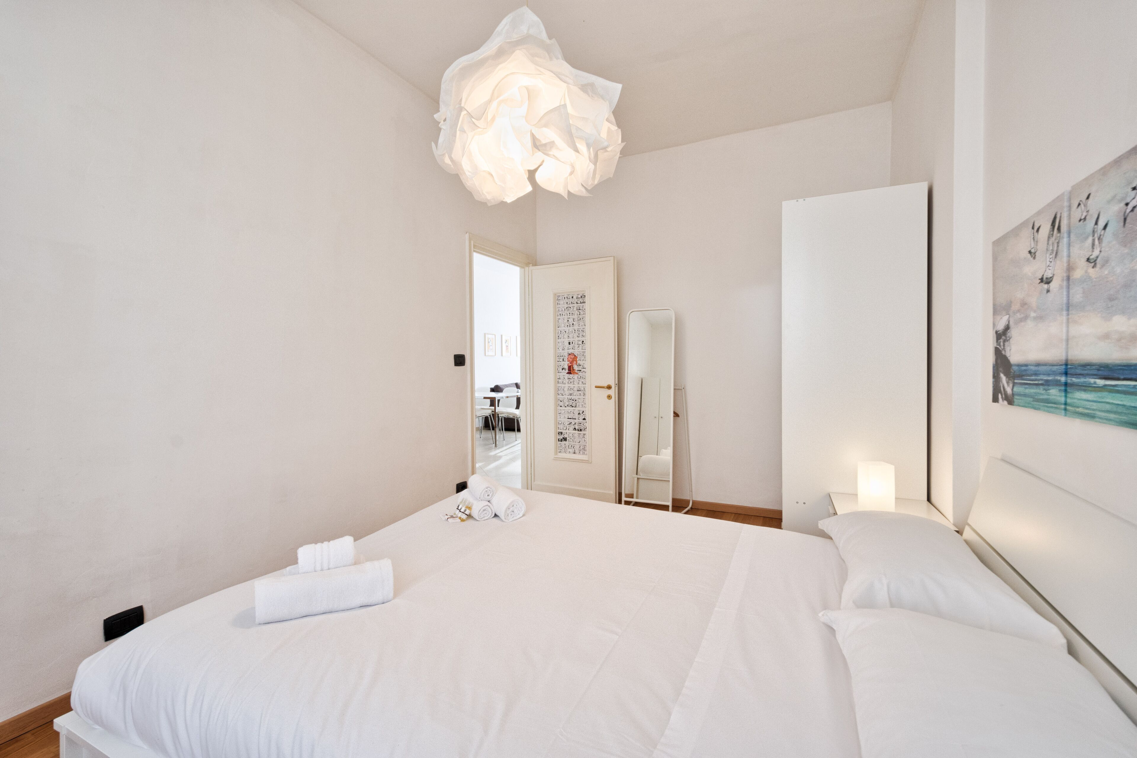 A hotel room with a large bed, a white ceiling light, a painting on the wall, and a door leading to another room.
