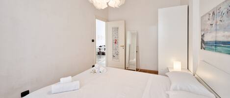 A hotel room with a large bed, a white ceiling light, a painting on the wall, and a door leading to another room.