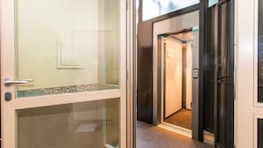 Entrada de hotel com porta de vidro, elevador e piso ladrilhado.