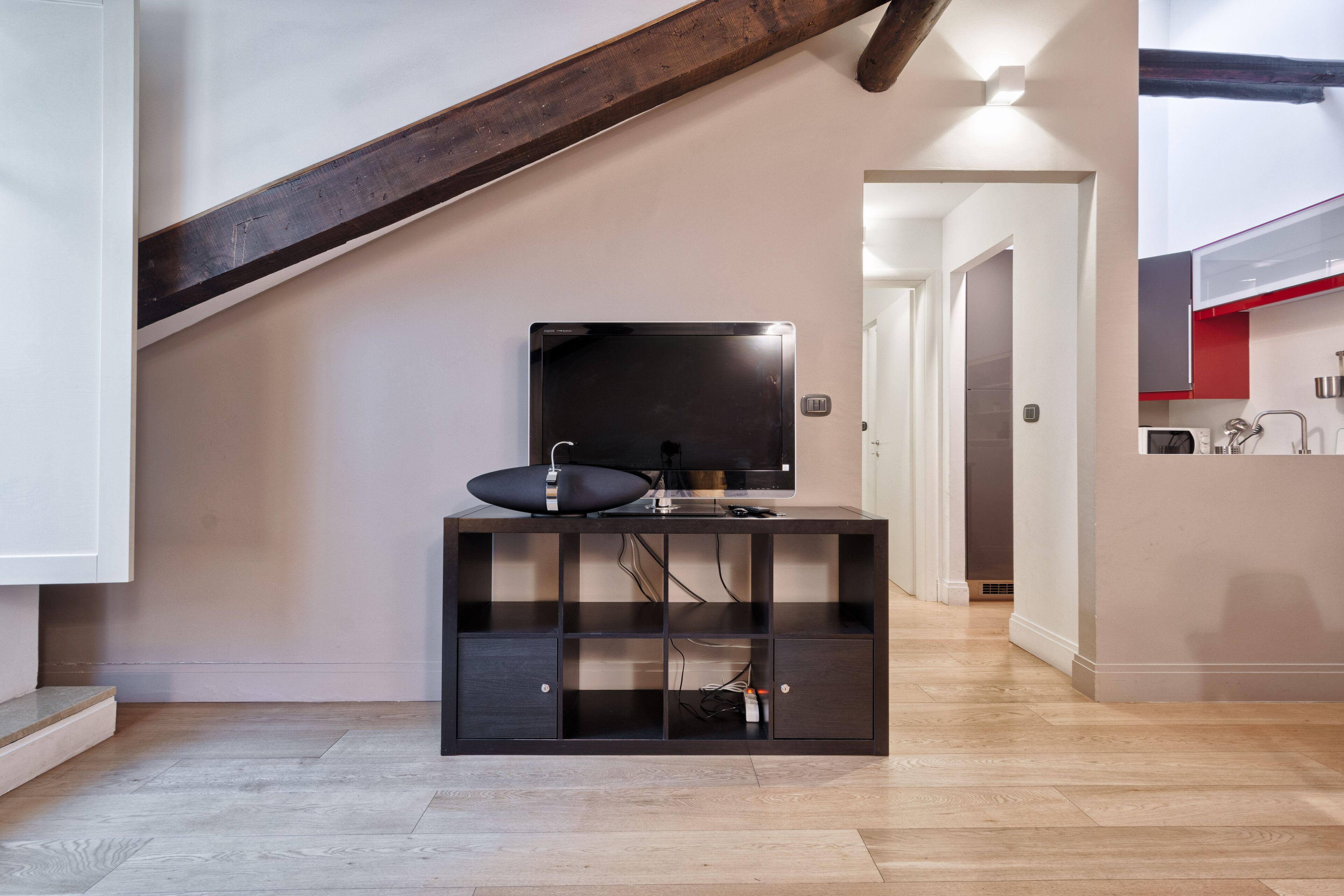 Ett modernt vardagsrum med en trappa i trä, en platt-TV på ett svart TV-bord och ett köksområde som syns genom en öppen dörr.