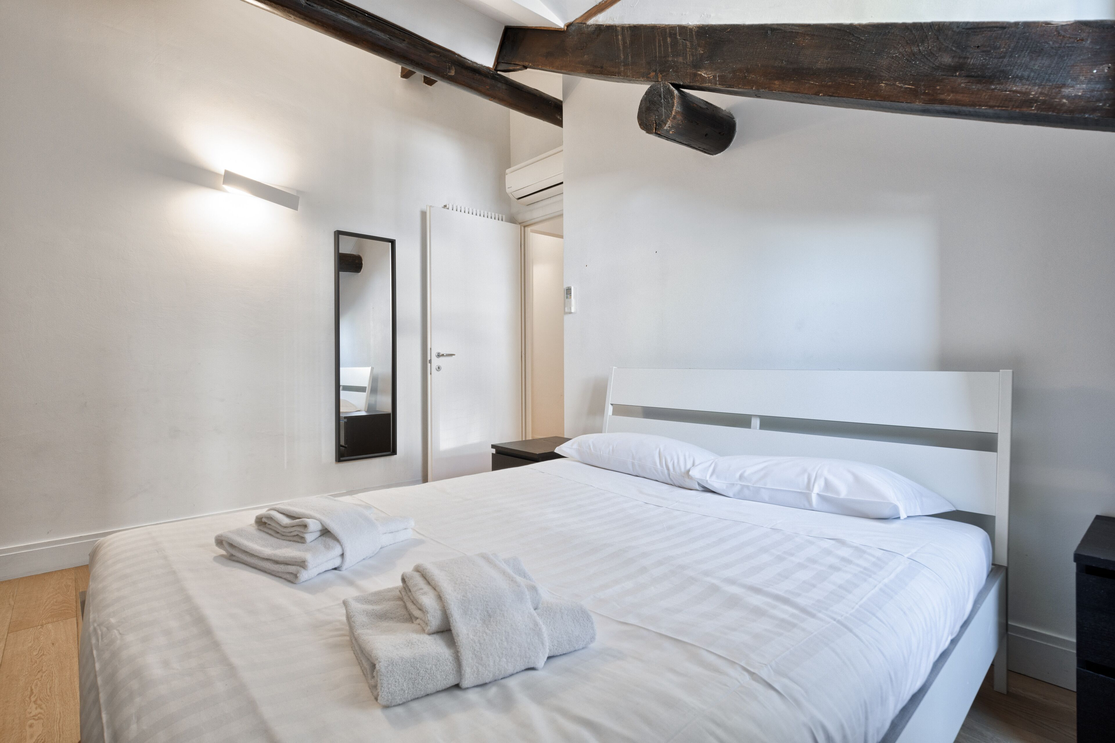A modern bedroom with a large bed, white linens, a wooden headboard, a nightstand, and a mirror on the wall.