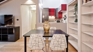 Ein moderner Essbereich mit einem dunklen Tisch und stuhlähnlichen Sitzgelegenheiten mit geometrischem Muster, einer großen Vasen-Zentralanordnung und einer Küche mit roten Schränken im Hintergrund.